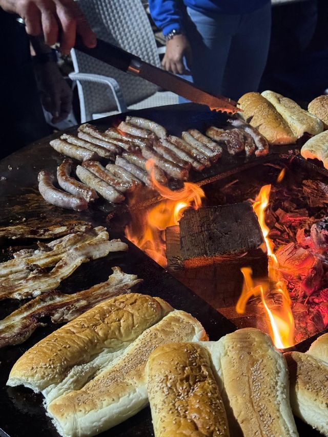 Primo piano di salsicce, pancetta e panini che cuociono su una grande griglia rotonda su fuoco a legna aperto.