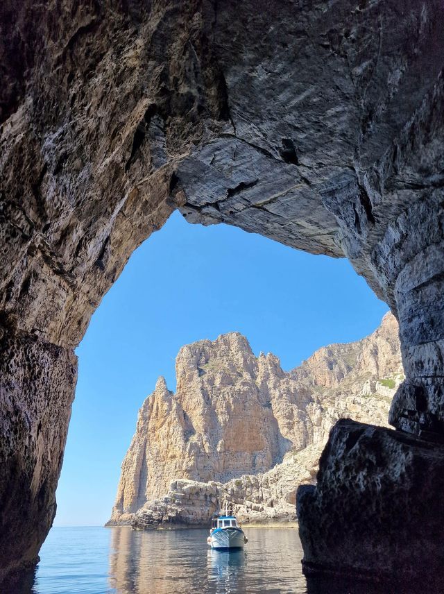 Una vista dall'interno di una grotta marina che guarda una piccola barca sull'acqua con scogliere rocciose sotto un cielo blu.