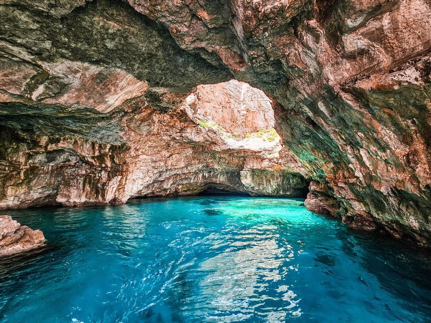 Vista dall'interno di un'ampia grotta marina con acqua limpida turchese. La luce del sole filtra attraverso un'apertura naturale nel soffitto roccioso.
