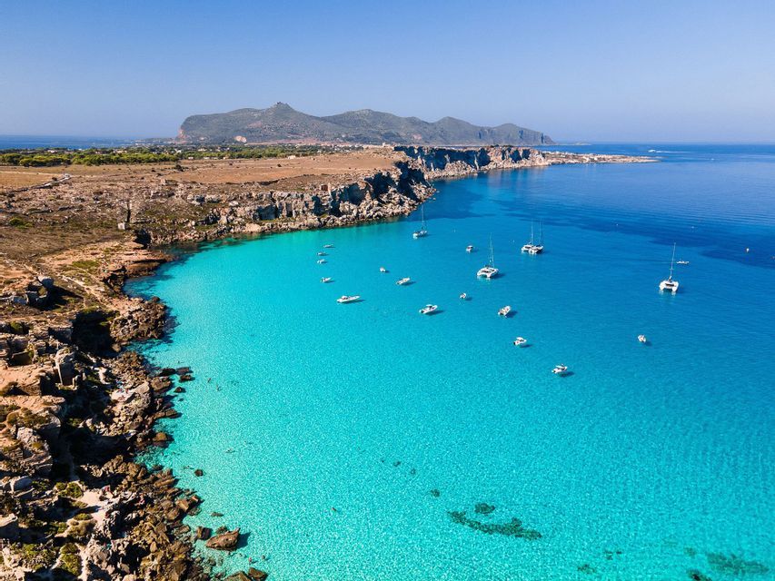 Vista aerea di barche a vela e catamarani ancorati in una baia turchese lungo una costa rocciosa.
