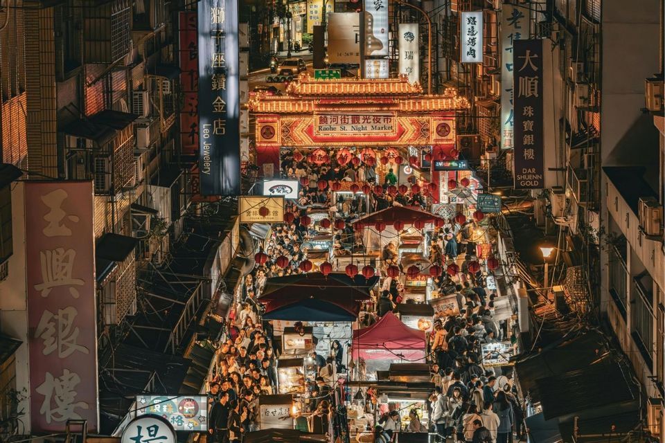 Eine Vogelperspektive auf einen belebten asiatischen Nachtmarkt, auf dem Menschen zwischen Ständen unter beleuchteten Schildern und roten Laternen umhergehen.