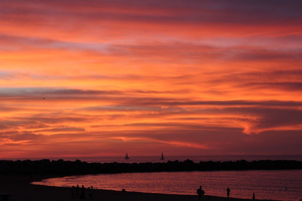 A vibrant orange sunset over the ocean, with silhouettes of people on the beach and sailboats on the water.