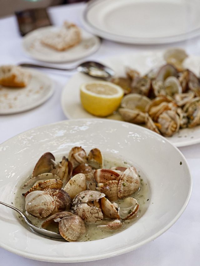 A white bowl of steamed clams in a broth is on a table with another plate of clams, bread, and a lemon wedge.