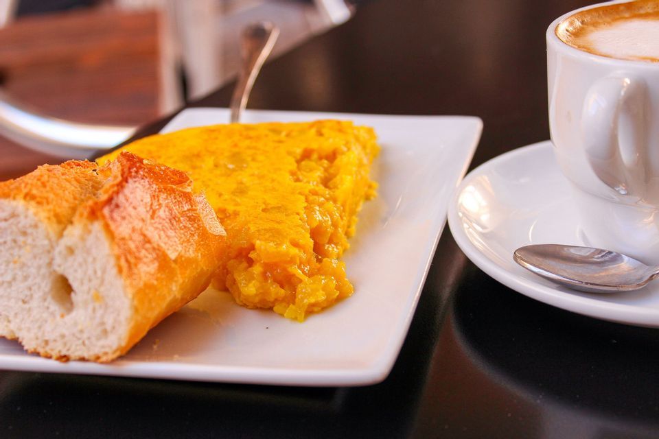 A close-up of a slice of Spanish omelette and a piece of bread on a white plate, next to a cup of cappuccino on a dark table.