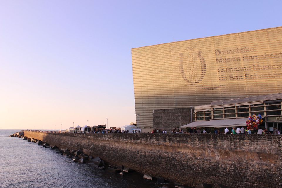 A modern building with a golden facade stands next to a stone sea wall where people are gathered by the water at dusk.
