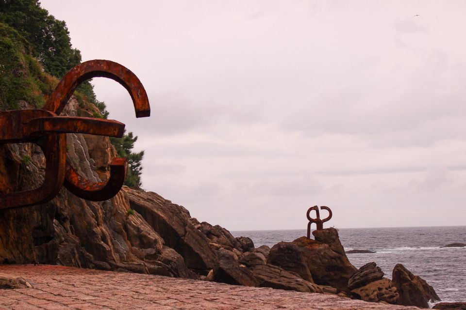 Two abstract, rusted metal sculptures are positioned on a rocky coast overlooking the sea under a cloudy sky.