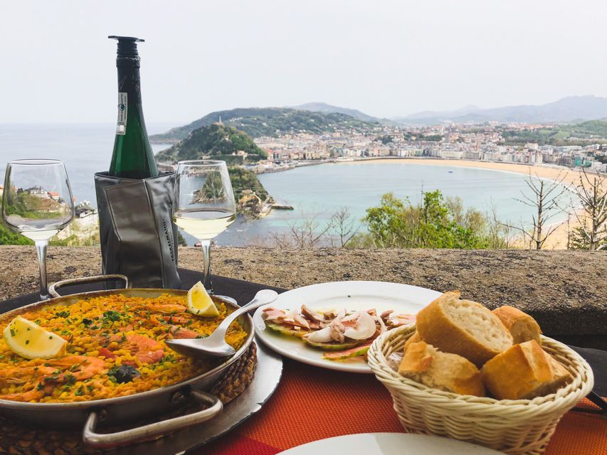 A meal of paella, wine, and tapas sits on a table overlooking a coastal city and a large, crescent-shaped beach.