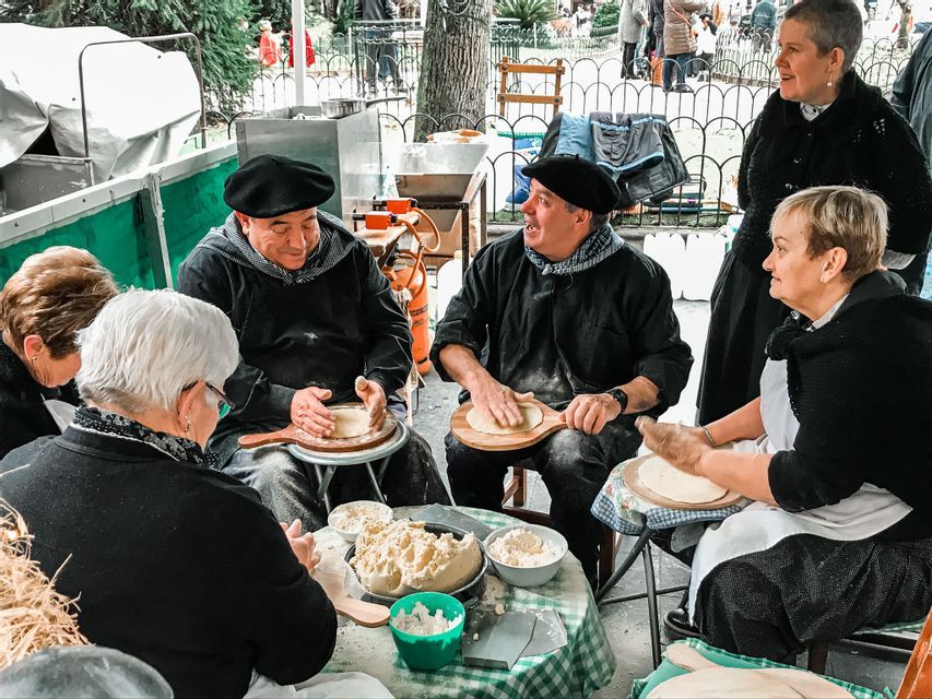 A group of people in traditional black berets and clothing prepare dough by hand on wooden paddles at an outdoor market.