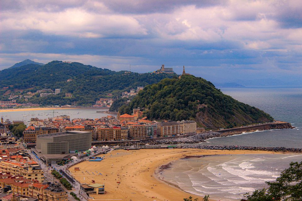 An elevated view of a coastal city with a large sandy beach, a bay, and a lush green hill by the sea under a cloudy sky.