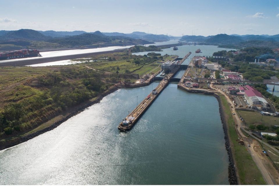 Una vista aérea de un gran sistema de esclusas de un canal, con varios buques de carga navegando por la vía fluvial, rodeado de colinas verdes e infraestructura.