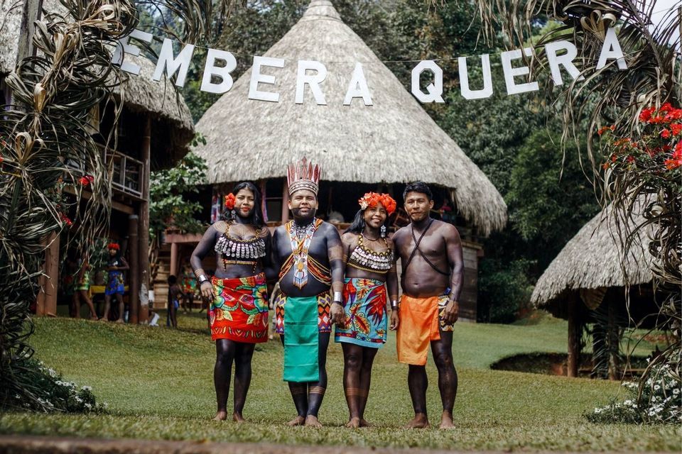 Cuatro personas Embera Quera con vestimenta tradicional y pintura corporal posan para una foto frente a cabañas con techos de paja.