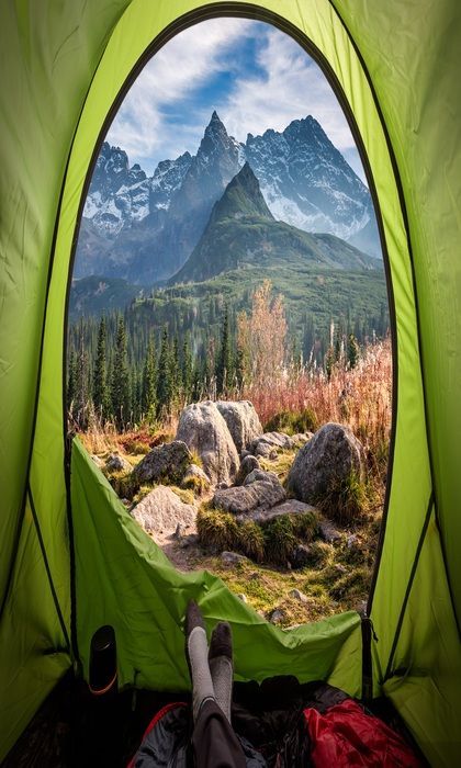 Una vista in prima persona dall'interno di una tenda verde, che si affaccia su una valle boscosa e montagne frastagliate e innevate.
