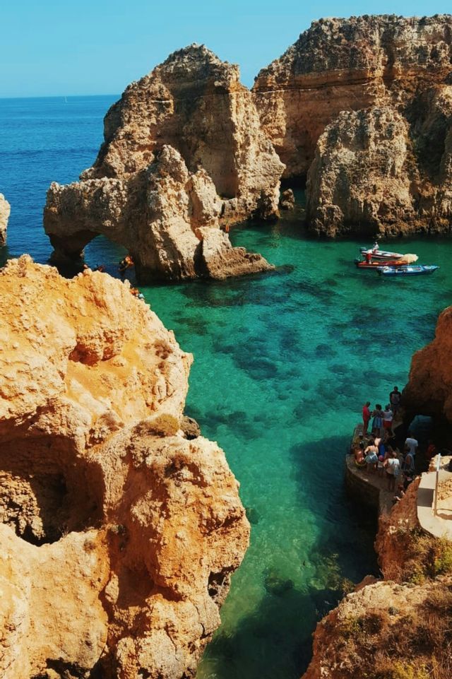 A WeRoad group trip gathers on a stone platform by a turquoise cove, surrounded by large rock formations and a sea arch.