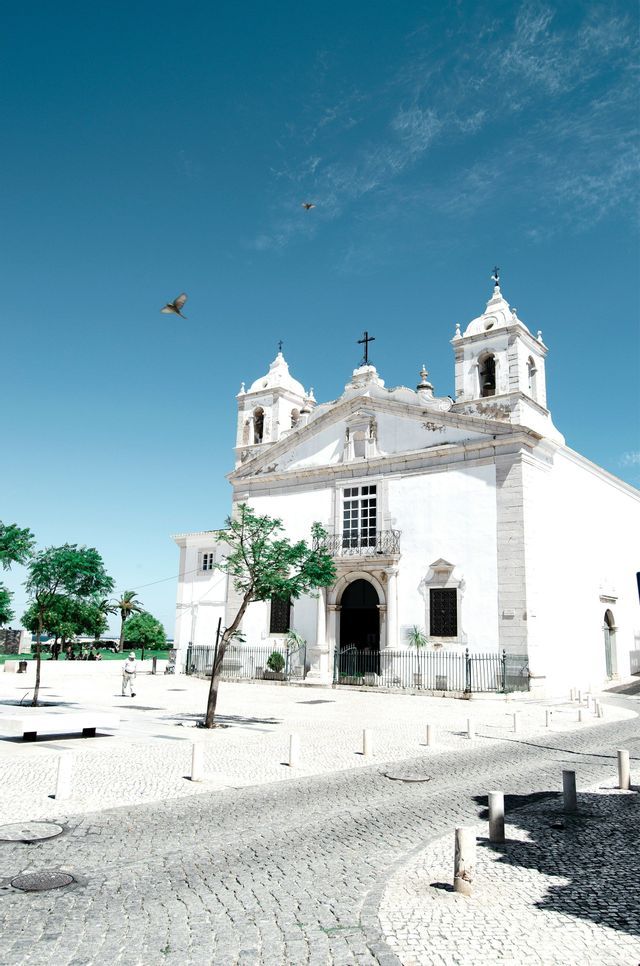 A white church with two bell towers stands in a cobblestone plaza under a clear blue sky with birds flying overhead.