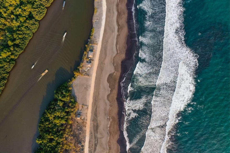 Eine Luftaufnahme eines Flusses mit Booten neben einem Sandstrand, der an den türkisfarbenen Ozean mit weißen Wellen grenzt.