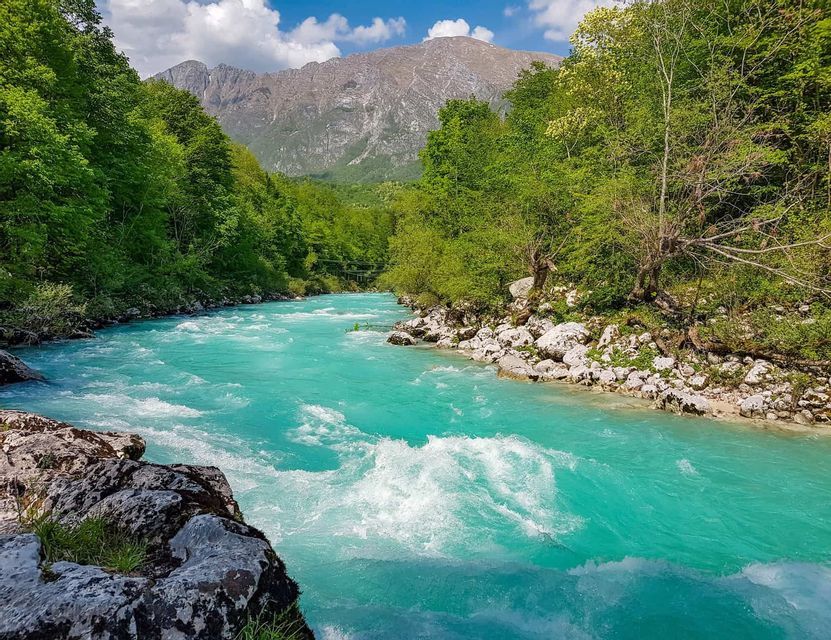 A turquoise river with white water rapids flows through a lush green valley with a mountain in the background.
