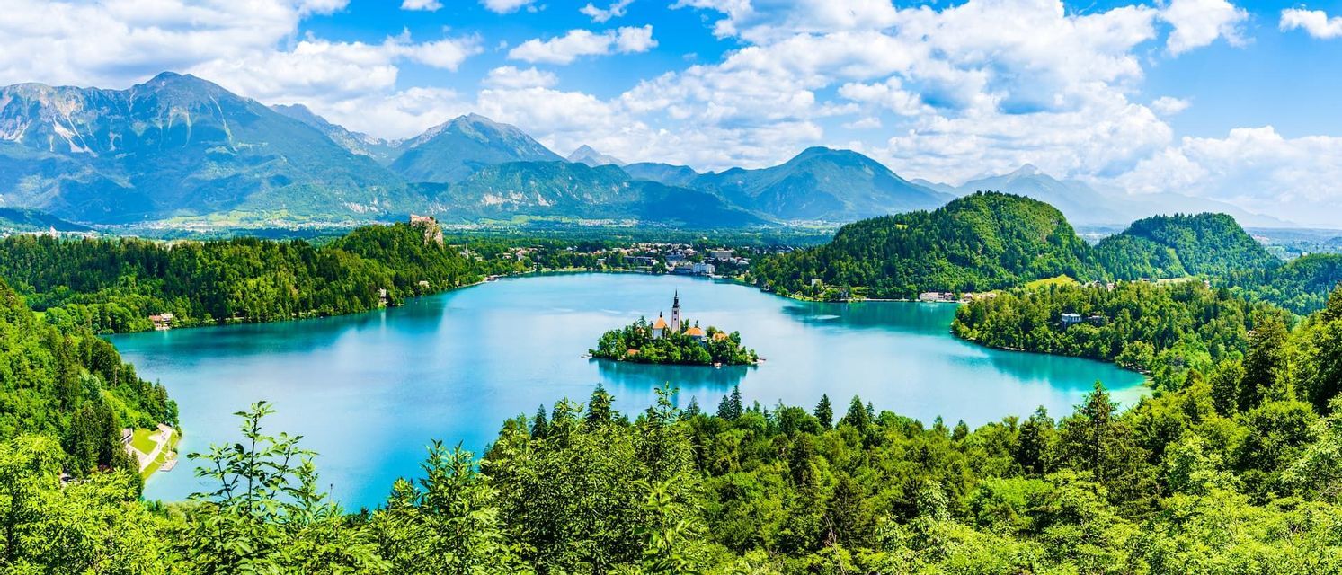 Panoramic view of a small island with a church in the middle of a blue lake, surrounded by green forests and mountains.