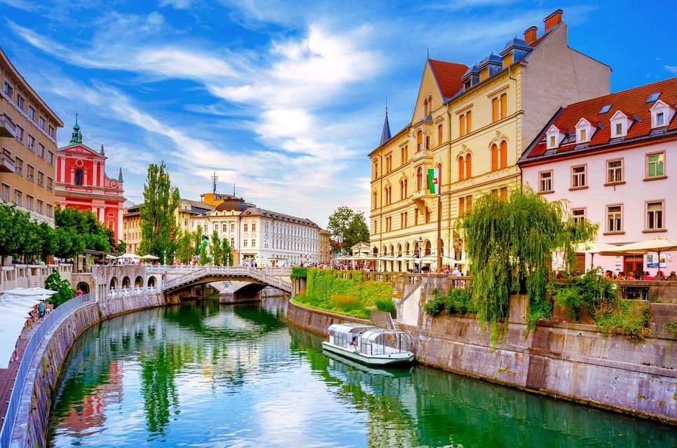 A river canal winds through a city with historic buildings, a stone bridge, and a tour boat on a sunny day.