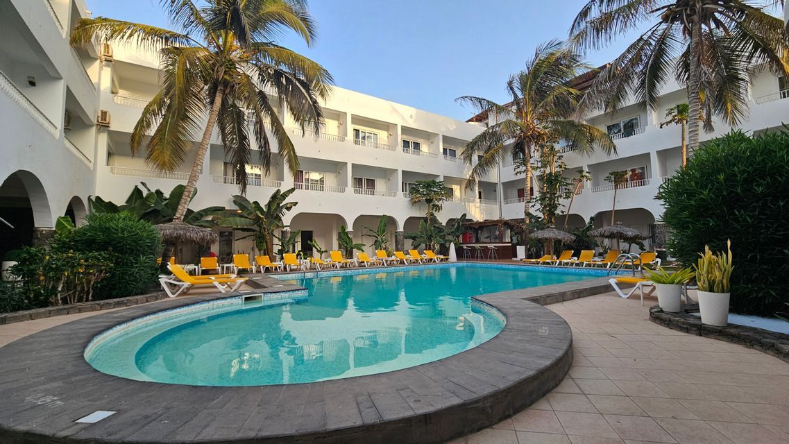 Ein türkisfarbenes Schwimmbecken, umgeben von gelben Liegestühlen und Palmen im Innenhof eines weißen Hotelgebäudes.