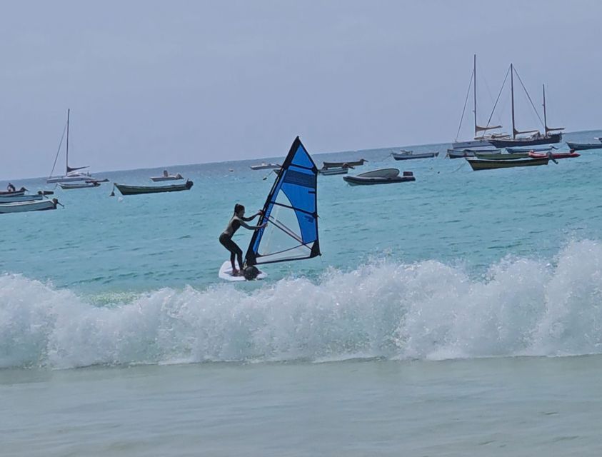 Una donna che fa windsurf su acqua turchese con una grande onda in primo piano e diverse barche ancorate sullo sfondo.