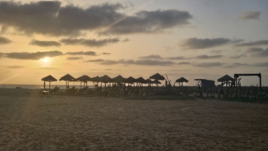 Una fila di ombrelloni di paglia e lettini da spiaggia stagliati sulla sabbia durante un tramonto nuvoloso sull'oceano.