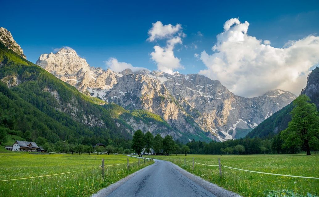 Una strada lastricata attraversa una rigogliosa valle verde verso una catena montuosa innevata e soleggiata sotto un cielo blu con nuvole.