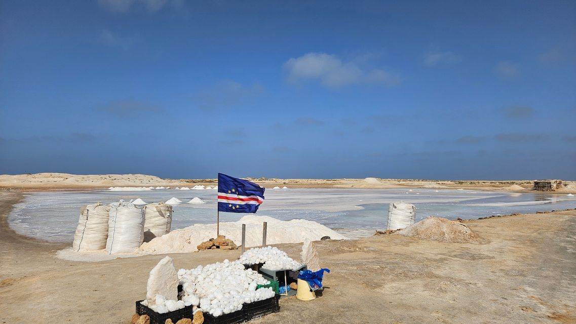 La bandiera di Capo Verde sventola sulle saline con cumuli e sacchi di sale raccolto sotto un cielo azzurro.