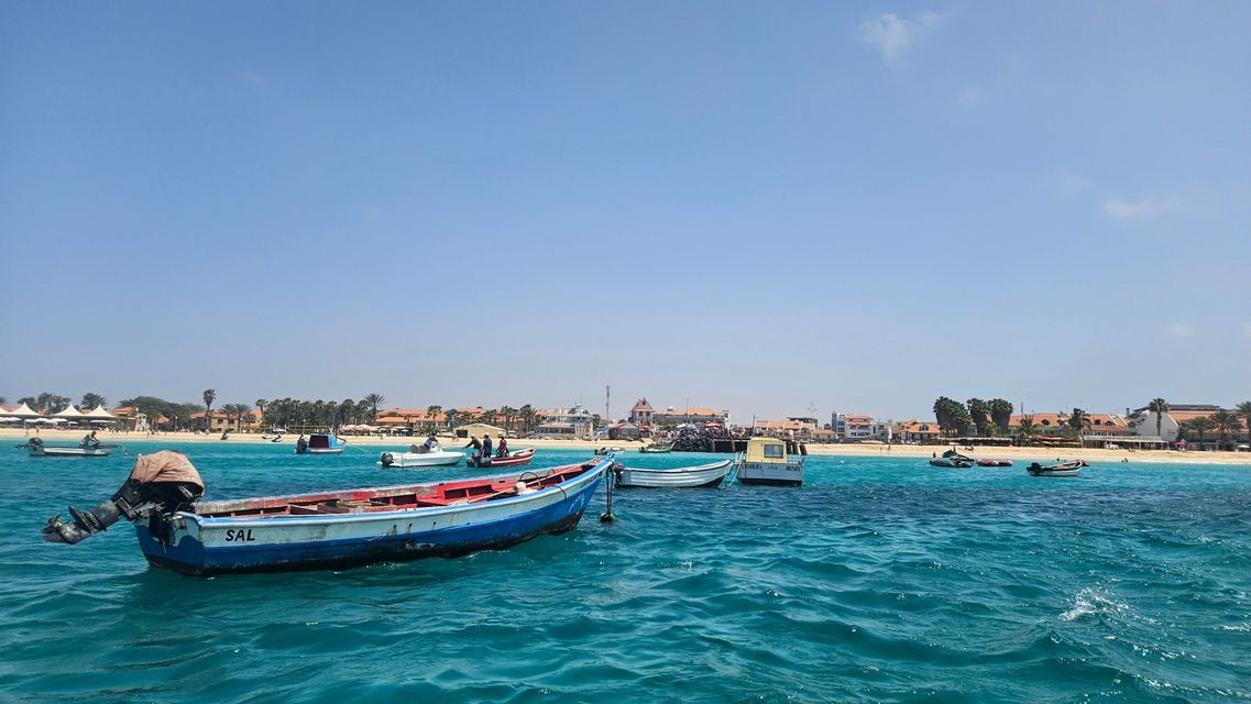 Una barca da pesca blu e rossa con la scritta 'SAL' sulla fiancata galleggia su acqua turchese, con altre barche e una località balneare sullo sfondo.