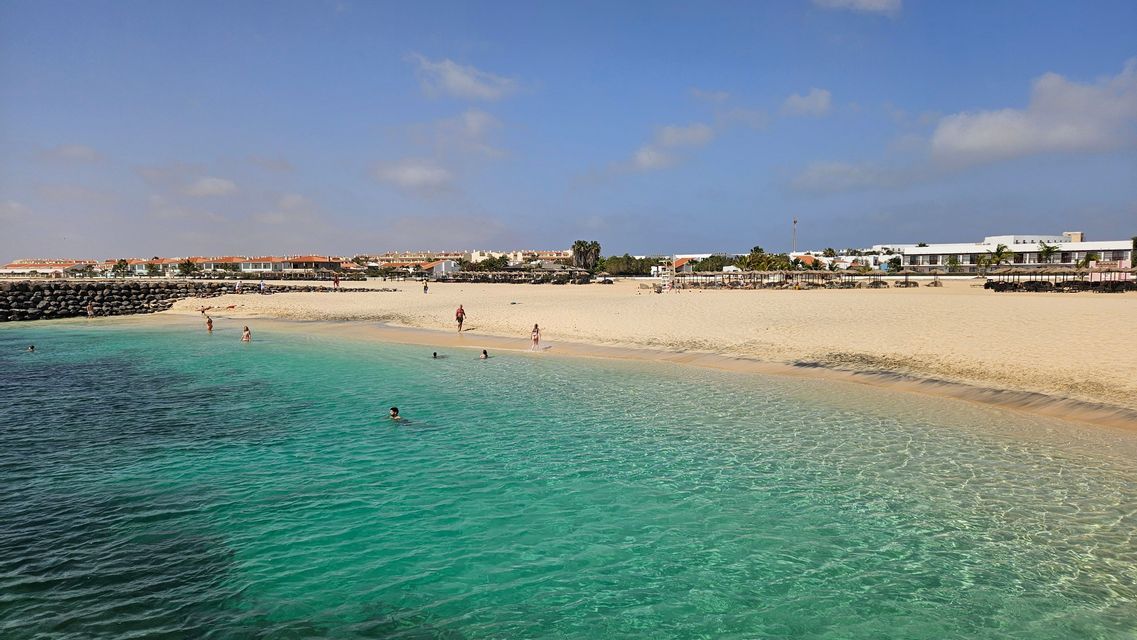 Spiaggia di sabbia bianca con bagnanti in acqua turchese cristallina e città costiera sullo sfondo
