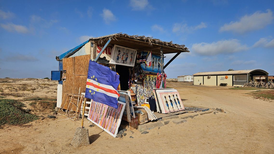 Un puesto de recuerdos en un paisaje arenoso exhibe arte y artesanía local, con una bandera de Cabo Verde ondeando en primer plano.