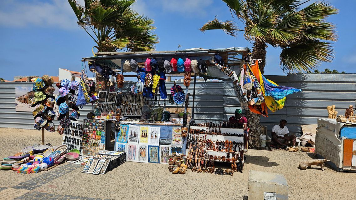 Un colorido puesto de recuerdos al aire libre exhibe artesanías, sombreros y pinturas hechas a mano a la venta bajo palmeras en un día soleado.
