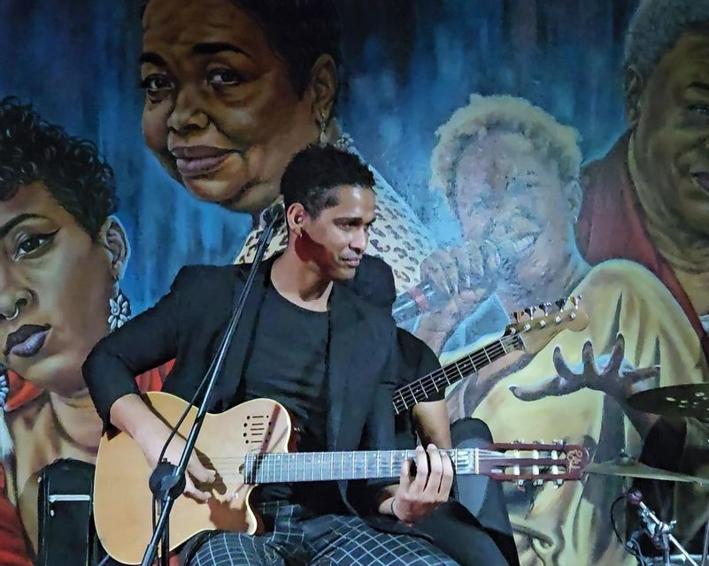 Un musicista con una giacca nera siede e suona una chitarra acustica davanti a un grande murale che raffigura diversi ritratti di cantanti.