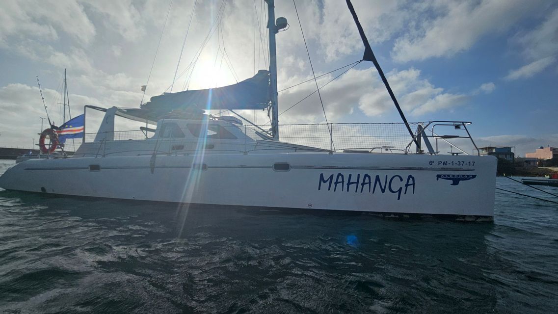 Un catamarano bianco con la bandiera di Capo Verde galleggia sull'acqua, retroilluminato dal sole splendente in un cielo nuvoloso.