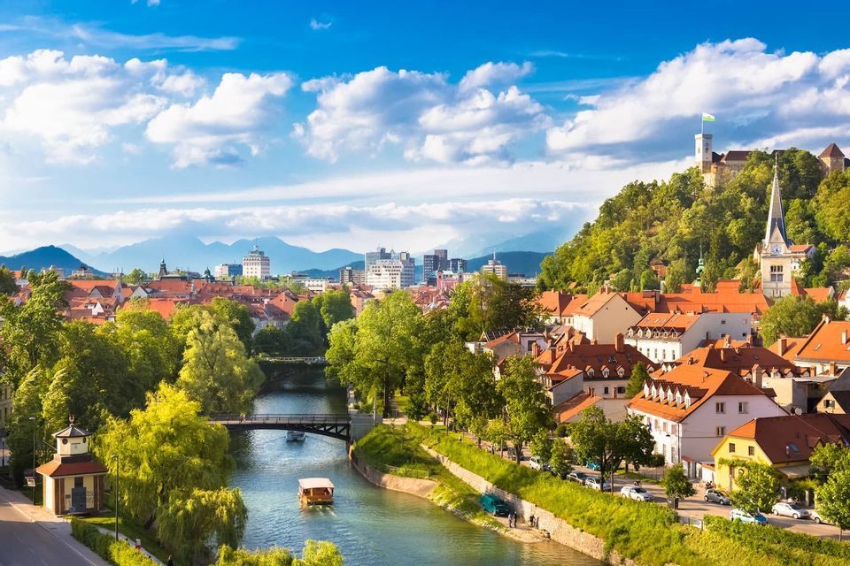 Eine Stadtlandschaft mit roten Ziegeldächern und üppig grünen Bäumen entlang eines Flusses, mit einer Burg auf einem Hügel und Bergen im Hintergrund.