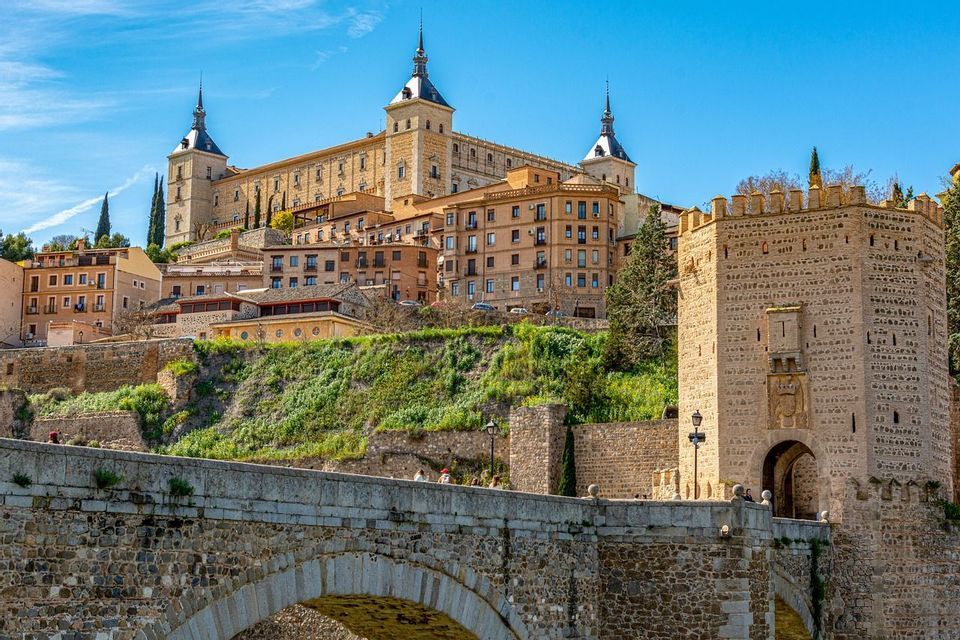 An arched stone bridge leads to a fortified gate and a historic city built on a green hill, with a large palace on top.