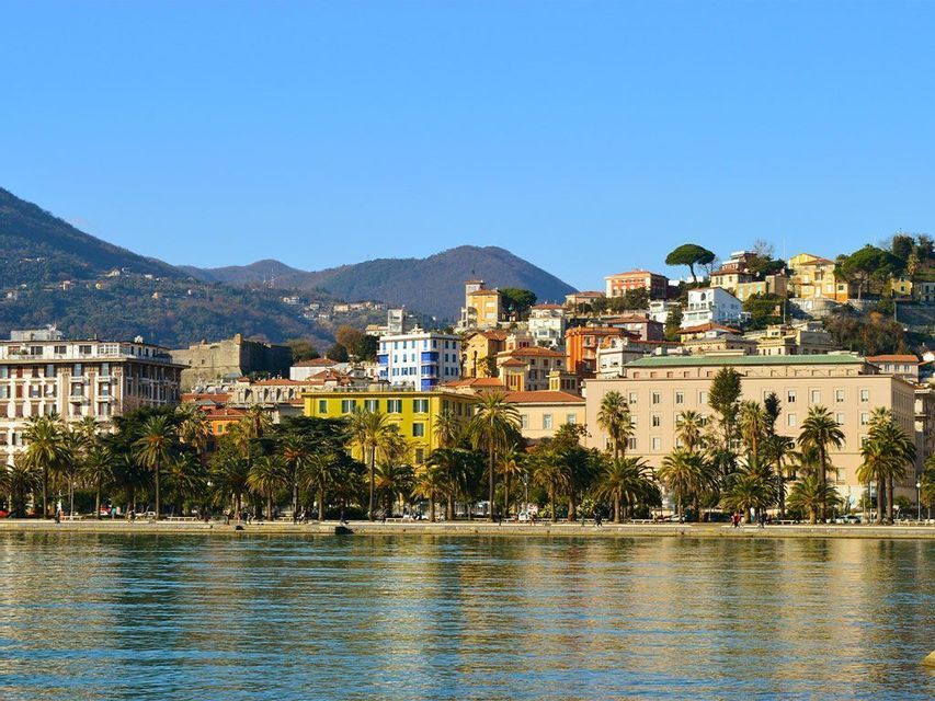Una vista sul lungomare di una città costiera con edifici colorati e palme, con verdi colline sullo sfondo sotto un cielo azzurro chiaro.