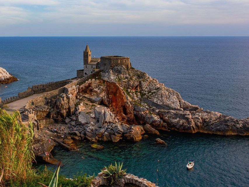 Une église en pierre avec un clocher se dresse sur une presqu'île rocheuse s'avançant dans la mer bleue, avec un petit bateau en contrebas.
