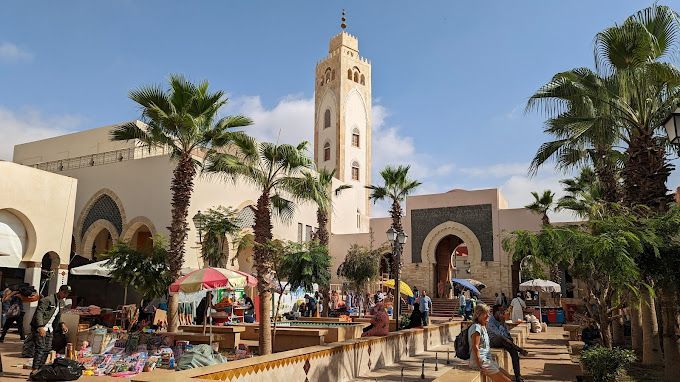 Une place de marché animée remplie de monde et de palmiers sous un ciel ensoleillé, avec un grand minaret en arrière-plan.