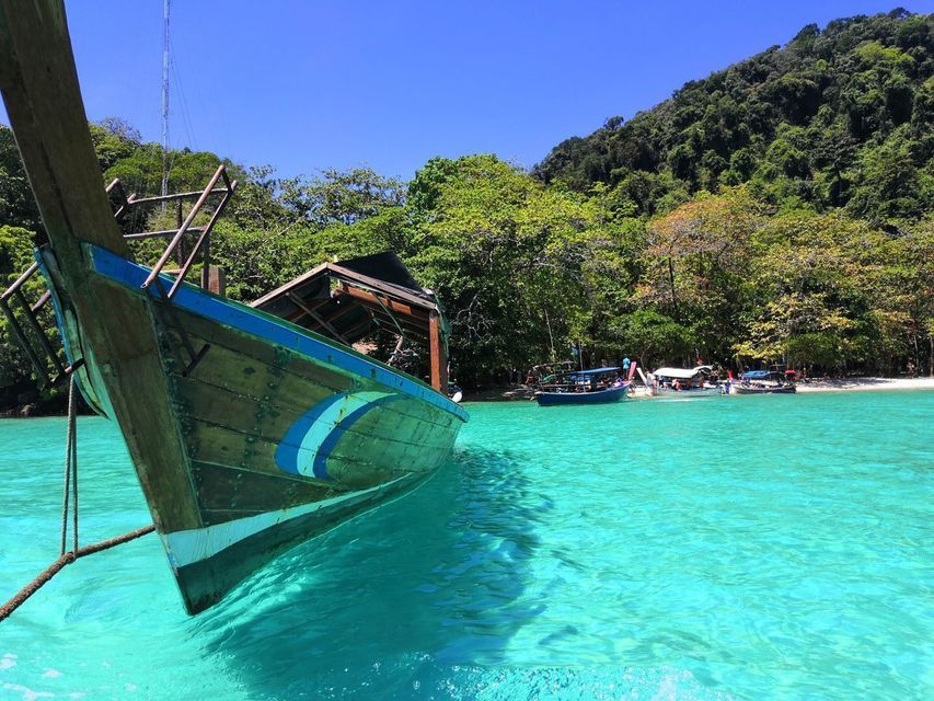 The bow of a traditional wooden boat moored in bright turquoise water, with a lush, forested island in the background under a clear sky.