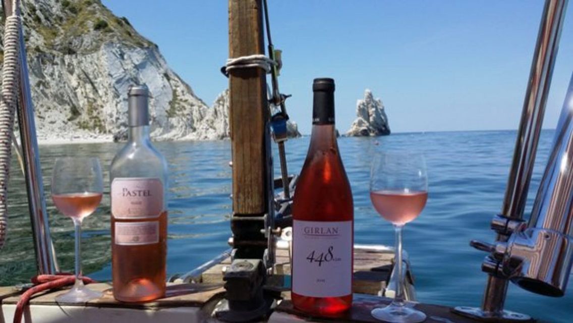 Due bottiglie di vino rosato e due bicchieri sul ponte di una barca a vela in mare, con scogliere sullo sfondo.