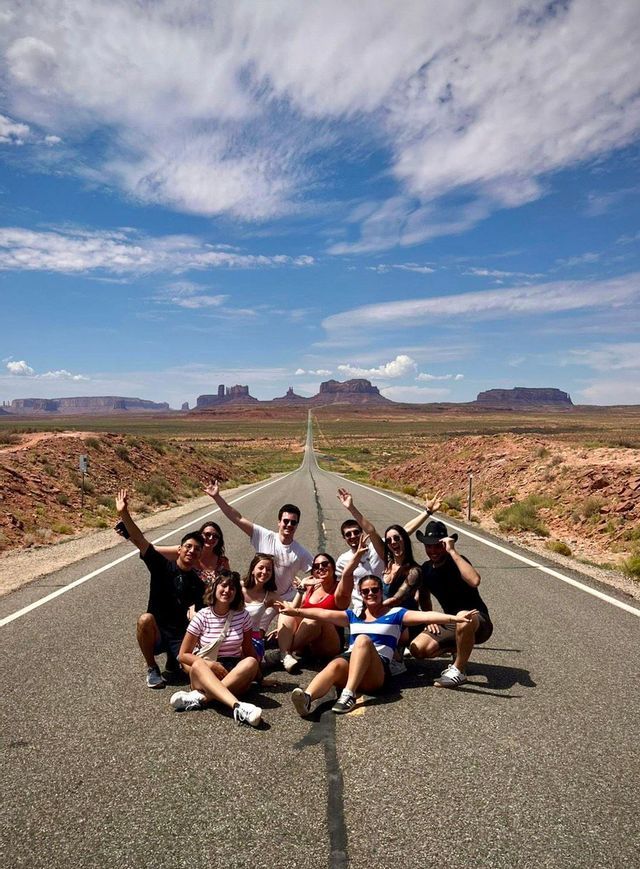 Un gruppo in viaggio con WeRoad posa al centro di una strada desertica che si estende verso lontane mesa sotto un cielo blu.
