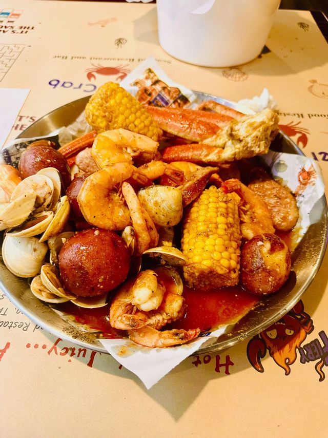 A close-up of a metal tray with a seafood boil of shrimp, crab legs, clams, corn, and potatoes covered in a red sauce.