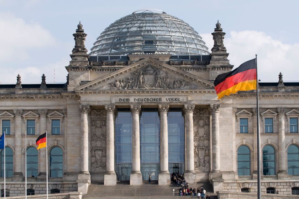 The facade of a neoclassical government building with a large glass dome, with German flags waving in the foreground.