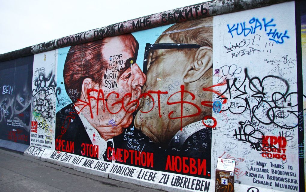 A mural on a concrete wall depicts two men in suits kissing, heavily covered by layers of graffiti and tags.