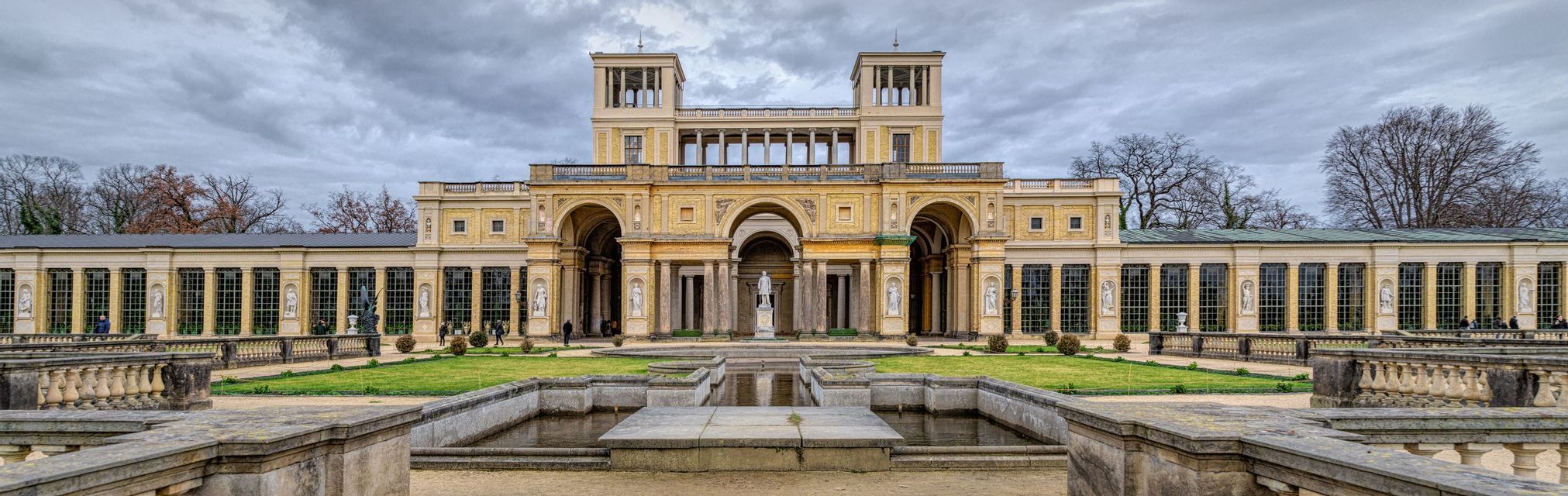 A panoramic view of an ornate classical palace with colonnades and a formal garden with fountains in the foreground under a cloudy sky.