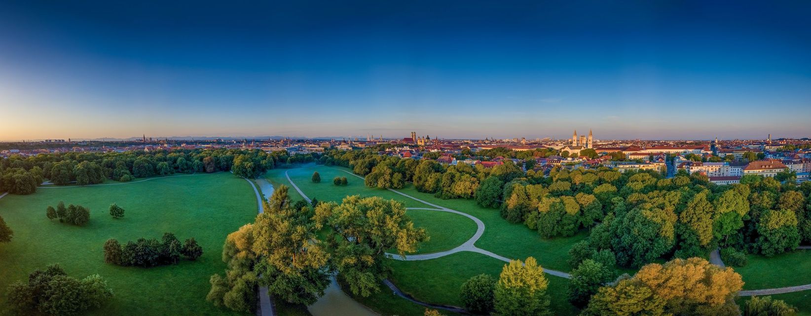 An aerial panoramic view of a city skyline next to a vast green park with trees and paths at sunset.