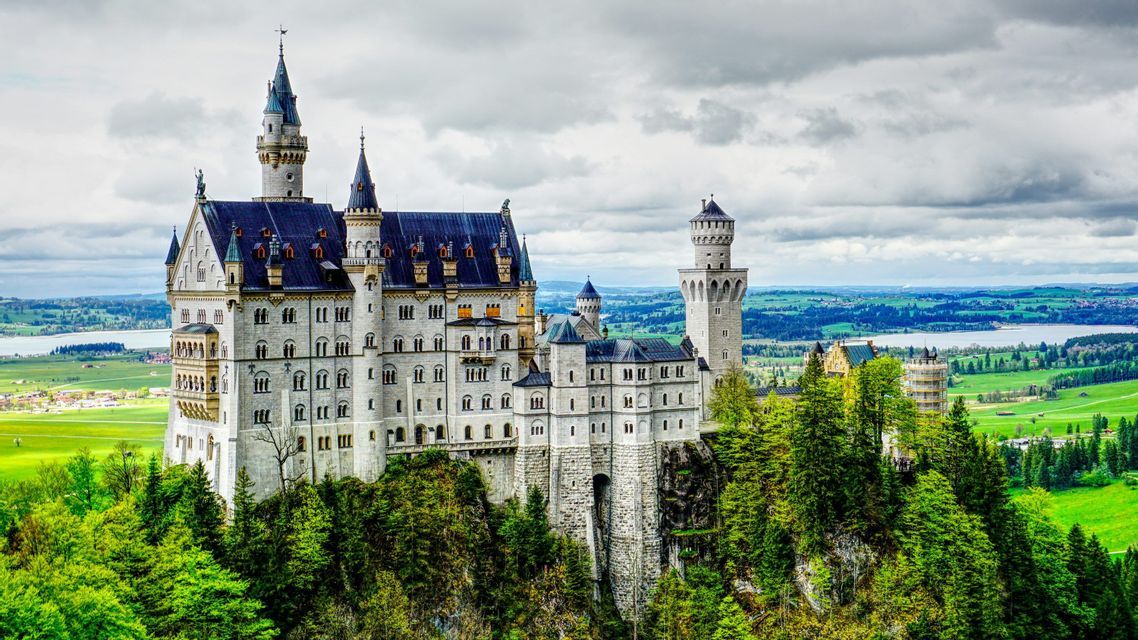A large castle with white walls and blue roofs stands on a forested hill, overlooking a vast green valley and a lake under a cloudy sky.