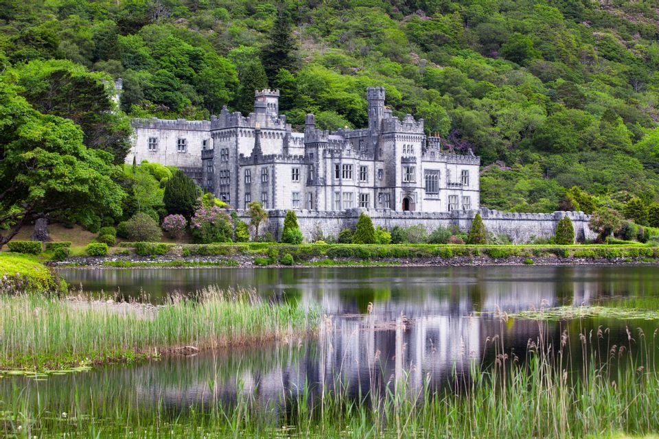 A large stone castle stands on the shore of a calm lake, its reflection visible in the water, with a lush green forest rising behind it.