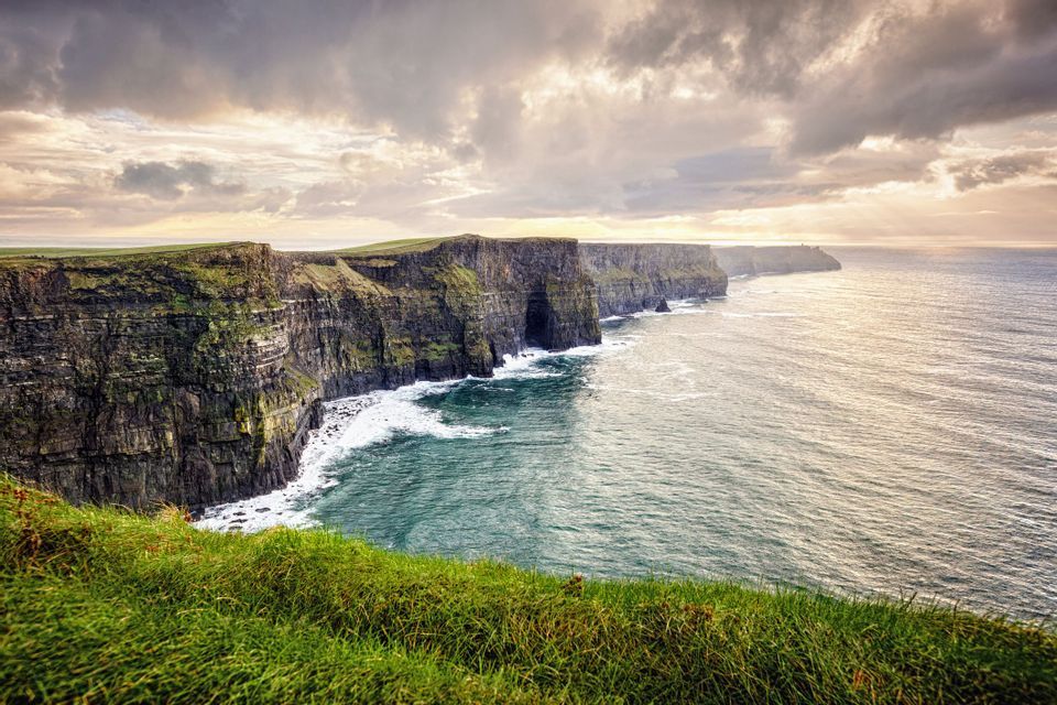 A coastline of steep, moss-covered cliffs meets the ocean, with white waves crashing at their base under a cloudy sky.