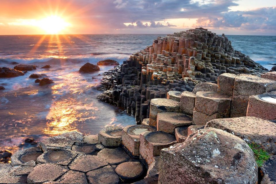 Hexagonal basalt rock columns line a coast as the sun sets over the ocean, its light reflecting on the waves.